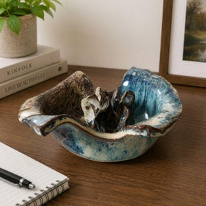 May include: A hand-formed ceramic bowl with an irregular, wavy rim. The exterior and interior feature a mottled glaze in shades of deep blue, white, and earthy brown. A sculptural, flower-like ceramic element is attached to the center of the bowl.