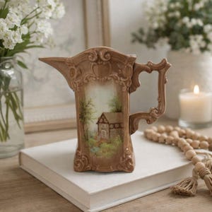 May include: A brown ceramic pitcher featuring an ornate, raised scrollwork border and a central painted scene of a cottage nestled among trees. The pitcher has a curved handle and a pointed spout, with a glossy finish and detailed relief molding.