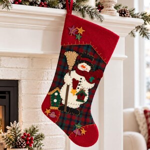 May include: A Christmas stocking featuring a red cuff and a dark green and red plaid body. The stocking displays a central snowman applique holding a broom, accompanied by small birdhouse motifs, star shapes, and heart accents in various colors.