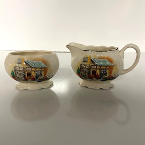 May include: A cream-colored sugar bowl and creamer set. Each piece features a hand-painted image of a quaint building with a blue roof, yellow trim, and red flowers. The creamer has a handle and a gold rim. The set is on a white surface.