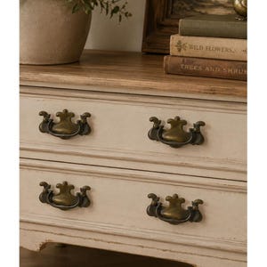 May include: A wooden dresser featuring two drawers with a distressed off-white finish and natural wood top. Each drawer is fitted with two ornate, antique brass-toned metal swing handles. The piece displays a weathered texture throughout.