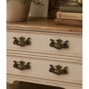 May include: A wooden dresser featuring two drawers with a distressed off-white finish and natural wood top. Each drawer is fitted with two ornate, antique brass-toned metal swing handles. The piece displays a weathered texture throughout.