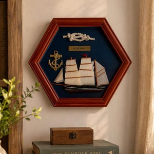 May include: A hexagonal wall display featuring a dark blue background inside a polished wooden frame. It includes a model sailing ship with white fabric sails, a gold-toned metal anchor, a rope knot, and a small plaque with the text NORWAY.