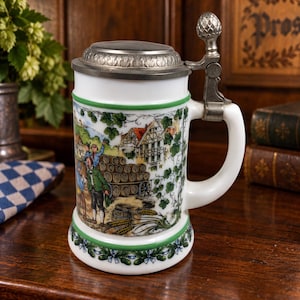 May include: A white ceramic beer stein featuring a colorful illustration of people near wooden barrels and a half-timbered house. The stein has a green rim, a decorative metal lid with a finial, and a sturdy handle. The base is adorned with a green floral garland pattern.