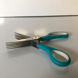 May include: A multi-blade herb scissor tool featuring five parallel stainless steel cutting blades and teal-colored handles with white interior grips. The design includes a central pivot point connecting the blades to the ergonomic handles.