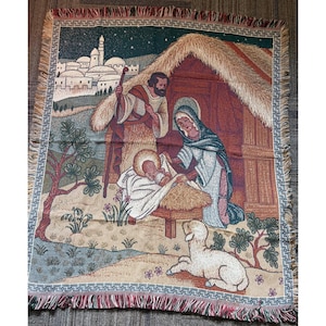 May include: A woven tapestry throw blanket featuring a nativity scene with a man, a woman, and a baby in a manger. The background shows a town with domed buildings under a starry night sky, a thatched-roof stable, trees, and a white lamb in the foreground.