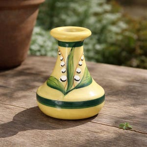 May include: A small, yellow ceramic vase with a bulbous base and a narrow neck. The vase is decorated with green leaves and white flower details. A wide green band encircles the vase near the base. The vase is sitting on a wooden surface.