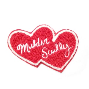 May include: Two red felt hearts sewn together with white embroidery. The hearts have the words "Mulder" and "Scully" written in white.