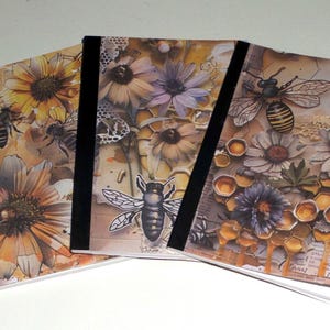 May include: Three notebooks with black spines, featuring floral and bee-themed designs. The covers showcase sunflowers, daisies, and honeycomb patterns in warm tones of yellow, orange, and brown, with bee illustrations.