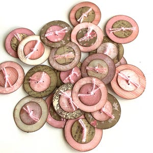 May include: A collection of layered, round buttons in shades of pink, cream, and brown. Each button features a smaller, central button secured with pink thread. Some have floral or map-like patterns.