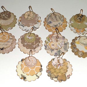 May include: A collection of ten handmade paper embellishments with a bee theme. Each has a scalloped edge, layered design, and a small metal clasp. The color palette includes beige, yellow, and brown, with floral and honeycomb patterns.