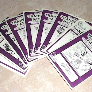 May include: A stack of eight purple and white iron-on transfer patterns for crafts. The patterns include designs for Easter, birthdays, and other occasions. The text on the patterns reads "Hot Iron Transfer Pattern".