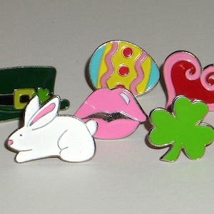May include: A collection of enamel pins featuring various designs. Includes a green leprechaun hat, a white bunny with pink ears, a colorful Easter egg, a pink heart, a pair of pink lips, and a green shamrock. Each pin has a silver border.