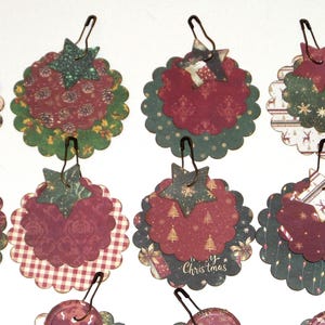 May include: A collection of circular paper tags with scalloped edges, each featuring festive patterns in red, green, and white. Each tag is topped with a small paper star and attached to a dark metal safety pin. Some tags display Merry Christmas text.
