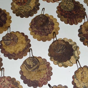 May include: A collection of handmade paper embellishments with a vintage aesthetic. Each piece features a scalloped edge, layered paper designs, and a small button or charm attached with a metal loop. The color palette includes shades of brown, beige, and burgundy.