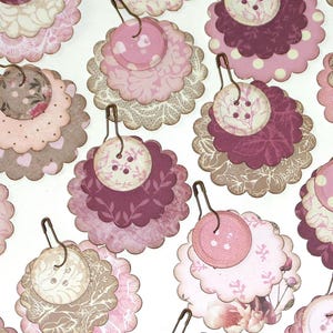 May include: A collection of layered paper embellishments in shades of pink, brown, and white. Each embellishment features a scalloped edge, a button, and a safety pin for attachment. The designs include floral, polka dot, and lace patterns.