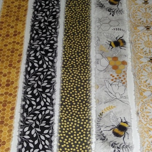 May include: Close-up of several fabric strips with bee and honeycomb patterns. The strips feature various designs, including a honeycomb pattern, black and white leaves, yellow dots, bees, and floral elements. The edges are frayed.