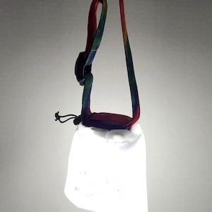 May include: A white, reflective drawstring bag with a colorful strap. The strap is a mix of red, green, and blue, and is attached to the bag with a black clasp. The bag is hanging against a gray background.