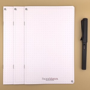 May include: A white, three-section notebook with a dotted grid pattern and a black pen. The notebook has a black logo that reads "FrontMatter" and "FRNTMTTR.COM".