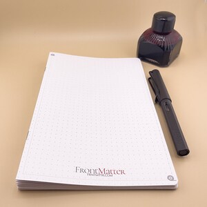 May include: A white notebook with a dotted grid pattern and the text "FrontMatter FRNTMTR.COM" printed on the cover. A black pen and a bottle of ink are on a beige surface next to the notebook.