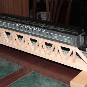 May include: A model train car labeled "New York Central" sits on a wooden trestle bridge. The train car is black and gray, and the bridge is made of light-colored wood. The scene is set on a table with a green and brown surface.