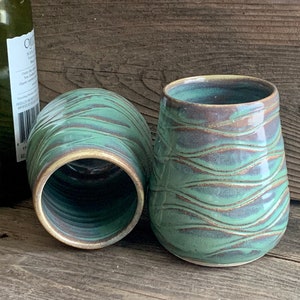Può includere: Due bicchieri da vino in ceramica verde e marrone con un motivo ondulato. I bicchieri sono su una superficie di legno.