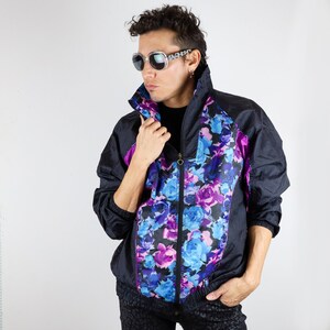 Puede incluir: Una chaqueta cortavientos negra con estampado floral en tonos azules, morados y rosas. La chaqueta tiene cierre de cremallera y cuello alto.