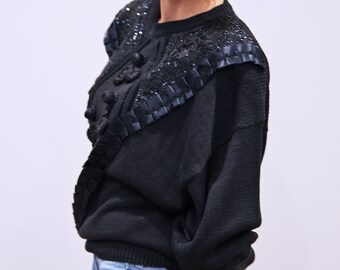 May include: Black sweater with a decorative black lace trim and embellishments. The sweater has a round neckline and long sleeves.