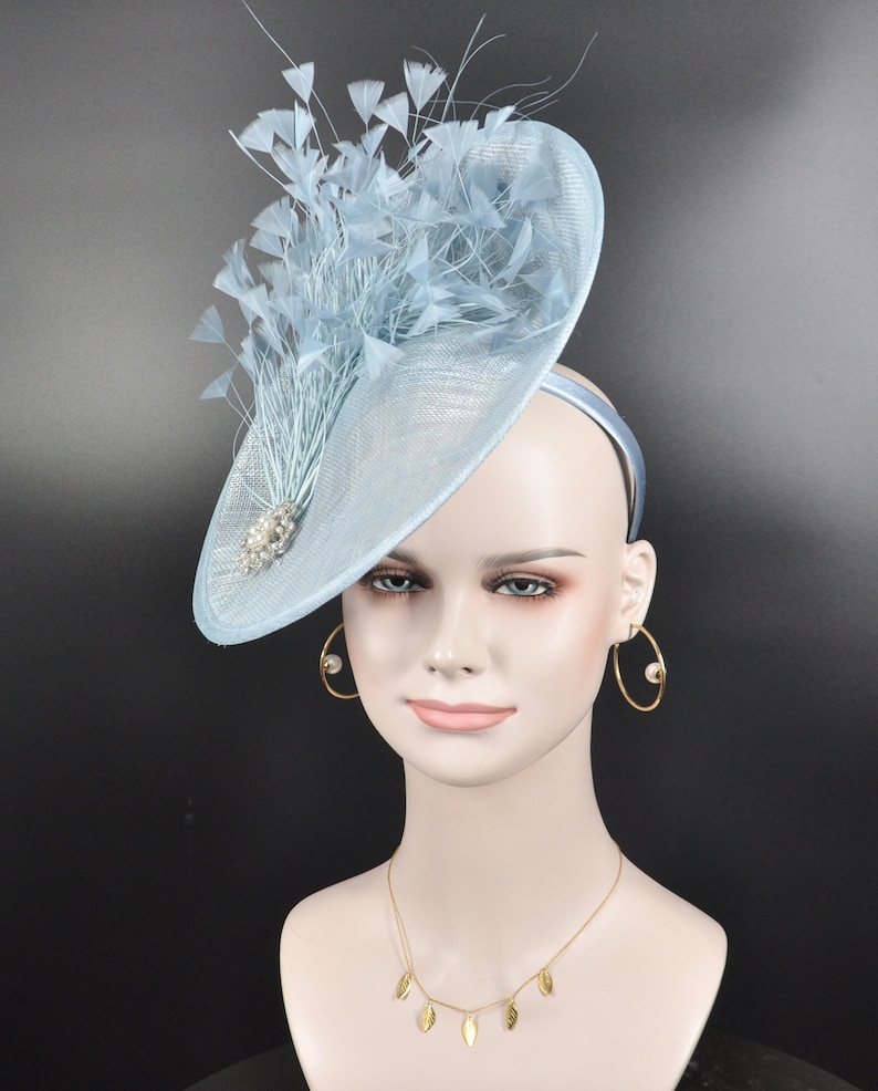 May include: A light blue fascinator hat with a large, round brim and a cluster of feathers on top. The hat is decorated with a small, sparkly embellishment.