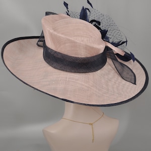 Pu&ograve; includere: Un cappello a tesa larga con una base in paglia rosa e un nastro e un bordo blu navy. Il cappello ha un velo in rete blu navy con un accento di piuma.
