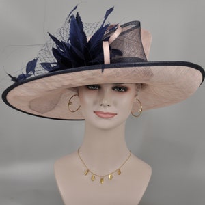 Pu&ograve; includere: Un cappello a tesa larga con una base in paglia rosa e un bordo blu navy. Il cappello &egrave; decorato con una piuma e una rete blu navy. Il cappello &egrave; indossato da un manichino con orecchini a cerchio dorati e una collana d'oro con ciondoli a forma di foglie.