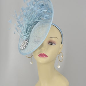 May include: A light blue fascinator with a large, curved brim and a feather accent. The fascinator is adorned with a sparkling rhinestone brooch.