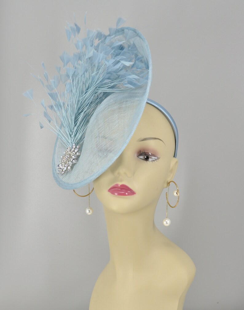 May include: A light blue fascinator with a large, curved brim and a feather accent. The fascinator is adorned with a sparkling rhinestone brooch.
