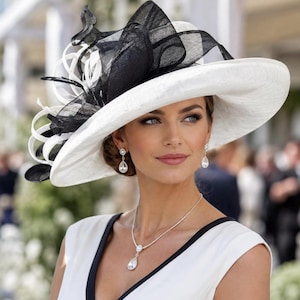 Puede incluir: Un sombrero blanco de ala ancha con un gran adorno decorativo en blanco y negro hecho de malla en capas y cintas en bucle. Se combina con un top blanco sin mangas con ribete negro, pendientes colgantes de plata y un collar de plata con colgante.