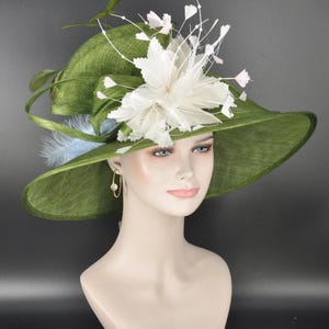 May include: A wide-brimmed olive green hat featuring a textured weave, adorned with a large white floral arrangement made of feathers and thin wire stems. A light blue feather accent is tucked under the brim, and gold hoop earrings are visible.