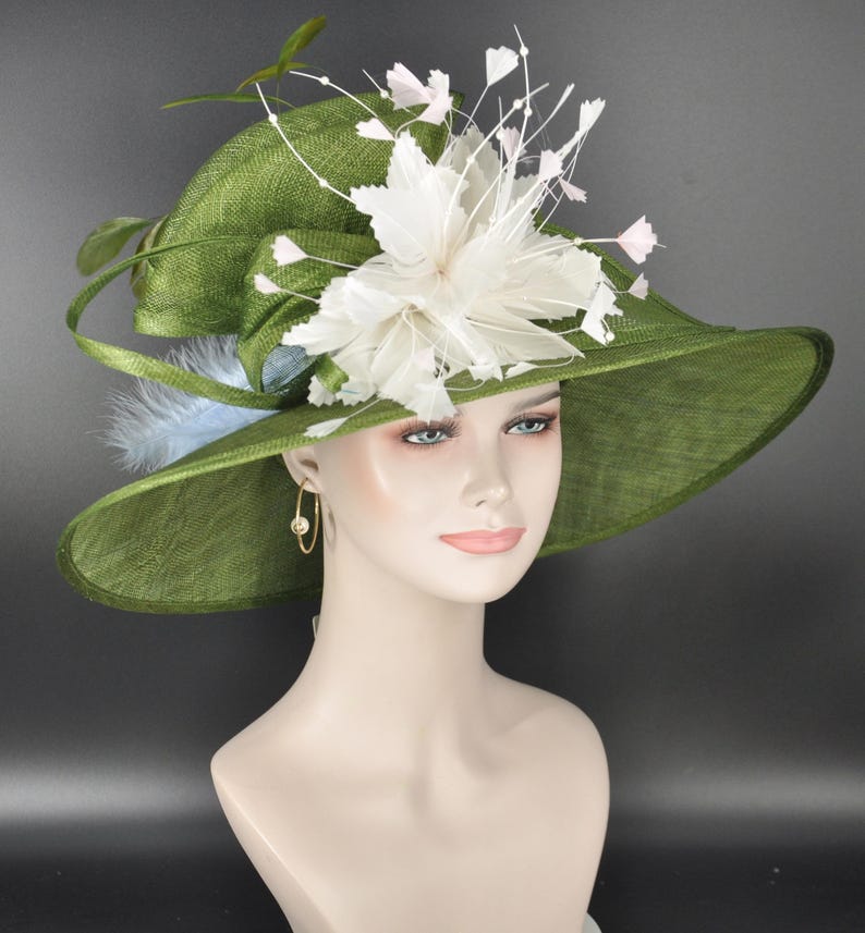 May include: A wide-brimmed olive green hat featuring a textured weave, adorned with a large white floral arrangement made of feathers and thin wire stems. A light blue feather accent is tucked under the brim, and gold hoop earrings are visible.