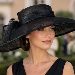 Puede incluir: Un sombrero negro de ala ancha confeccionado en un material tejido con textura. El sombrero presenta una copa estructurada rodeada por una cinta negra a juego y un gran lazo superpuesto en el lateral, logrando un aspecto formal.