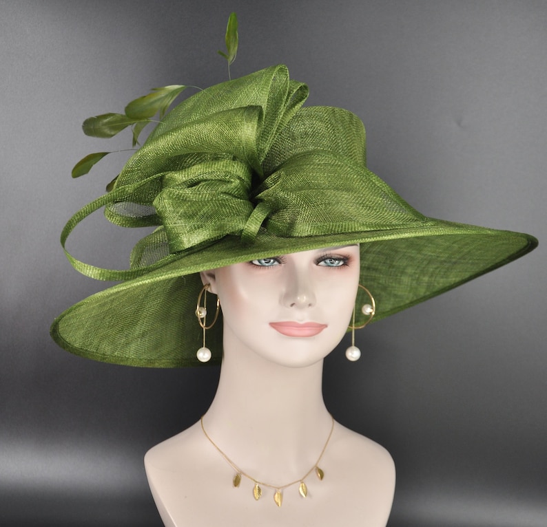 May include: An olive green wide-brimmed hat featuring a structured crown and a large, layered bow detail on the side. The hat is adorned with delicate green feathers extending from the top. The material has a visible woven texture throughout.