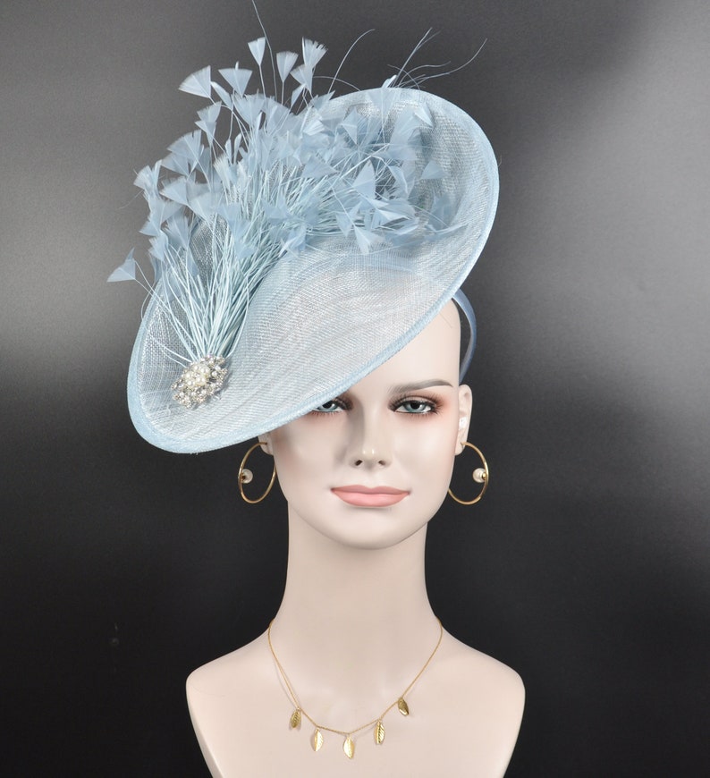 May include: A light blue fascinator hat with a large, wide brim and a cluster of feathers on top. The hat is decorated with a small, sparkly brooch.