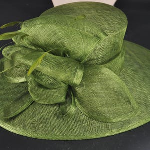 May include: A wide-brimmed olive green hat featuring a structured crown and a large, layered bow detail on the side. The hat is crafted from a textured, woven material and is accented with several thin, delicate feathers tucked into the bow.