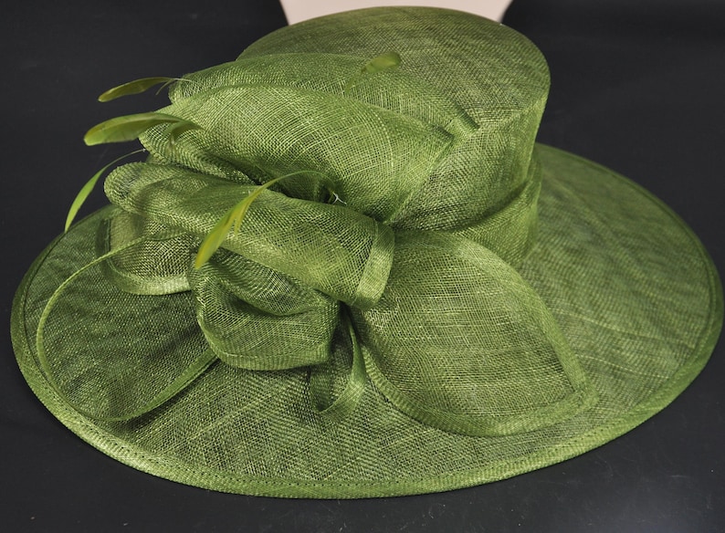 May include: A wide-brimmed olive green hat featuring a structured crown and a large, layered bow detail on the side. The hat is crafted from a textured, woven material and is accented with several thin, delicate feathers tucked into the bow.