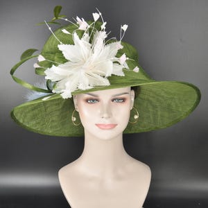 May include: A wide-brimmed olive green hat featuring a textured woven finish. The hat is adorned with a large, layered white floral embellishment made of feathers and delicate stems, positioned asymmetrically on the side of the crown.