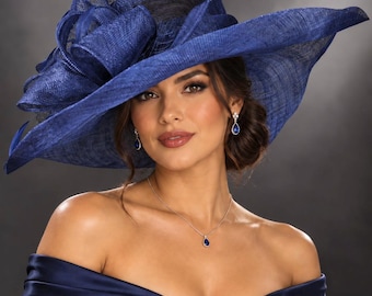 May include: A wide-brimmed navy blue hat featuring a large, structured bow detail made of textured fabric. The hat is paired with a matching navy blue off-the-shoulder dress, a silver necklace with a teardrop pendant, and teardrop earrings.