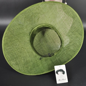 May include: A wide-brimmed olive green hat with a woven texture, featuring a circular crown and a black adjustable drawstring cord inside. A white rectangular tag is attached, displaying a graphic of a hat and the text Miss Race.