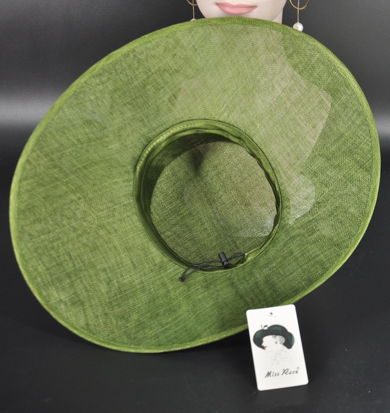 May include: A wide-brimmed olive green hat with a woven texture, featuring a circular crown and a black adjustable drawstring cord inside. A white rectangular tag is attached, displaying a graphic of a hat and the text Miss Race.