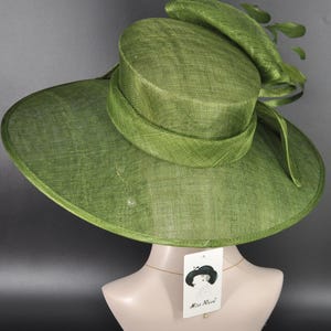 May include: A wide-brimmed olive green hat with a structured cylindrical crown and a matching band. The hat features a decorative bow element on the side with green leaves. A white rectangular tag hangs from the hat, displaying the text Miss Race.