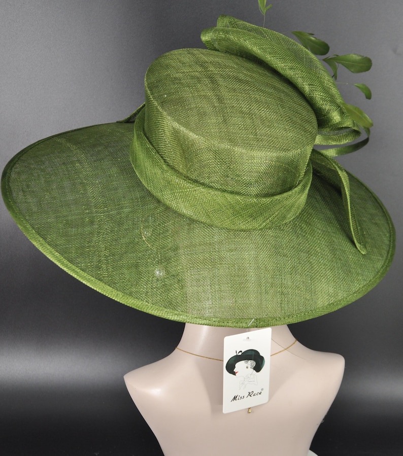 May include: A wide-brimmed olive green hat with a structured cylindrical crown and a matching band. The hat features a decorative bow element on the side with green leaves. A white rectangular tag hangs from the hat, displaying the text Miss Race.