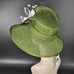 May include: A wide-brimmed olive green hat with a structured crown and a textured woven finish. The hat is adorned with a cluster of white floral accents and delicate sprigs on one side. A small white rectangular tag hangs from the back edge.