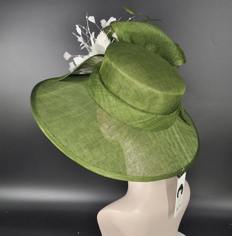 May include: A wide-brimmed olive green hat with a structured crown and a textured woven finish. The hat is adorned with a cluster of white floral accents and delicate sprigs on one side. A small white rectangular tag hangs from the back edge.