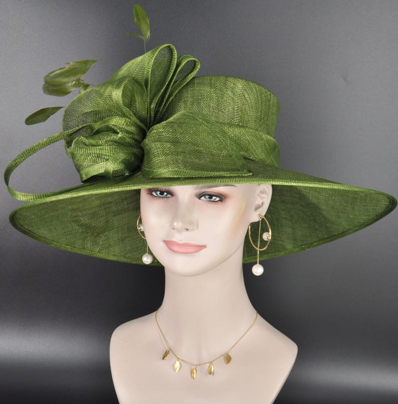 May include: An olive green wide-brimmed hat featuring a structured crown and a large, layered fabric bow accent on the side. The hat is displayed on a mannequin head wearing gold-toned drop earrings with white pearls and a gold leaf-motif necklace.